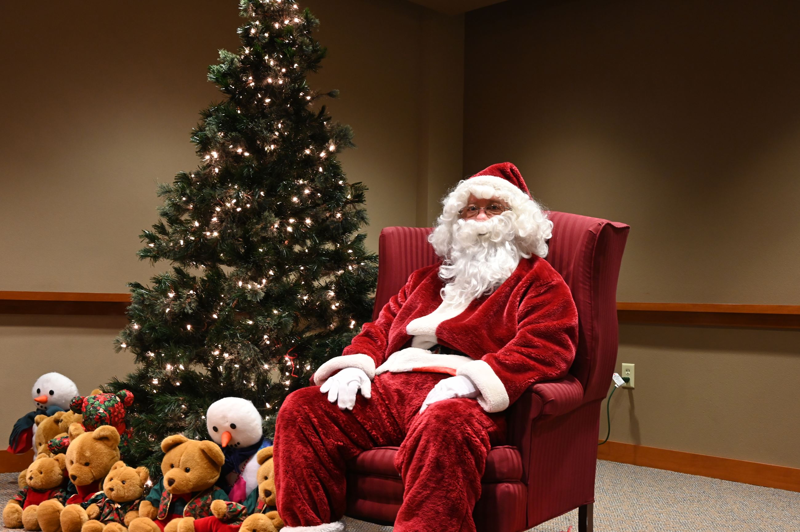 Santa Claus sitting on a red chair next to a decorated tree with many stuffed animals