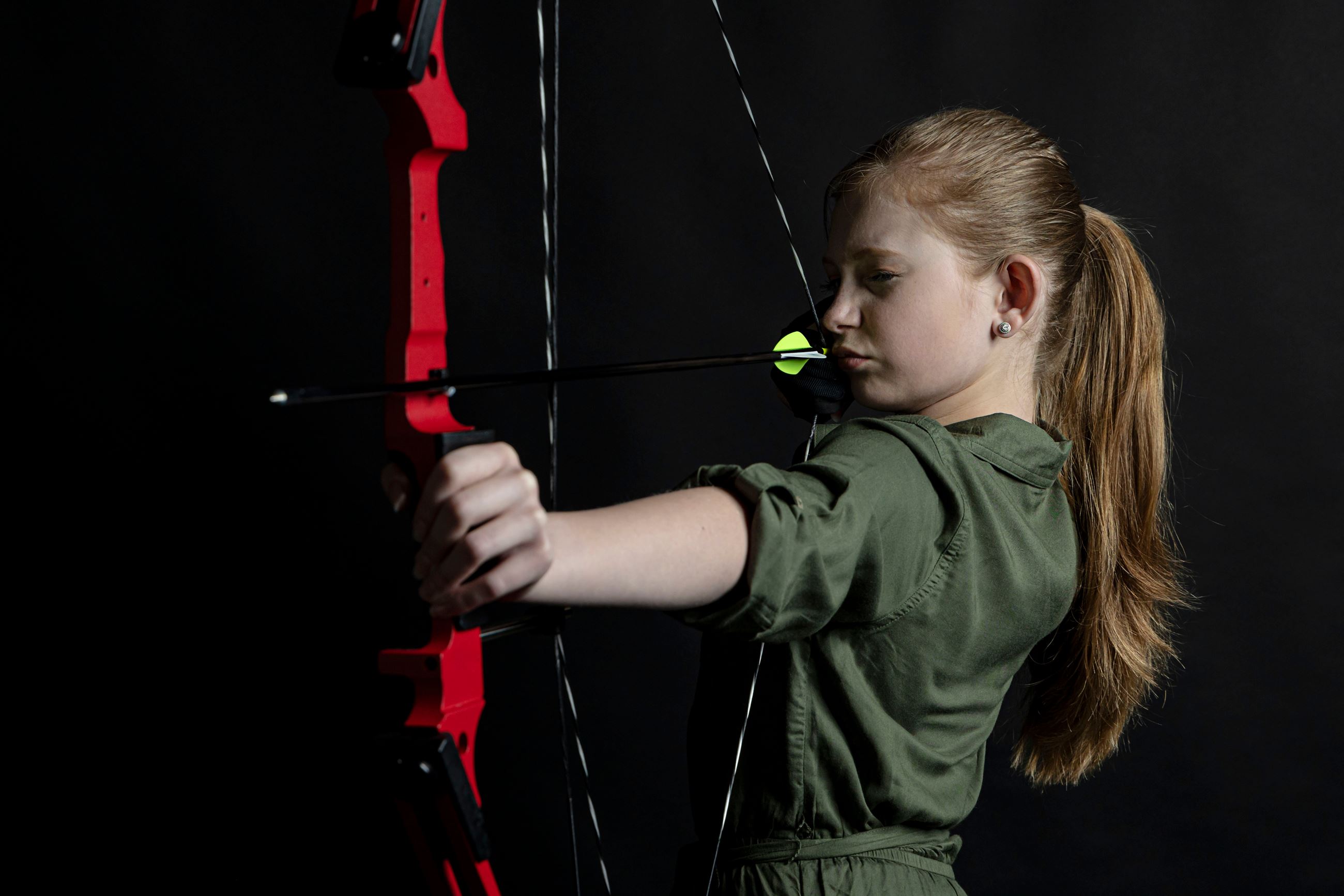 Young girl in the Archery program taking aim during a session against a dark background.